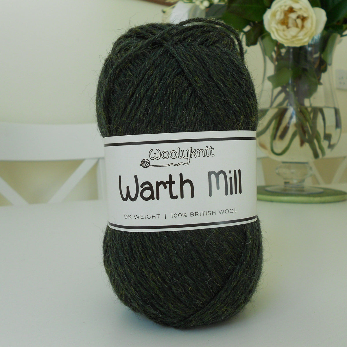A ball of Woolyknit Warth Mill yarn in a dark green colourway. It's sitting on a white table, with a vase of flowers in the background.